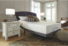 Colchón de Memory Foam tamaño Matrimonial sobre base ajustable oscura con cabecera tapizada capitoné, en recámara con buró blanco, lámparas y sillón de mimbre - Estylo House Muebleria