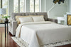Sofá Cama Queen Storrow extendido como cama con cubrecama blanco acolchado, tapizado gris con reposabrazos rectos, en sala con mesa lateral, lámpara y tapete texturizado - Estylo House Muebleria